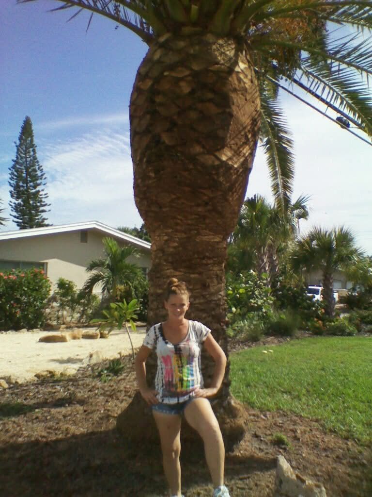 Jenniffer standing at the base of a tall date palm in a tidy front yard.
